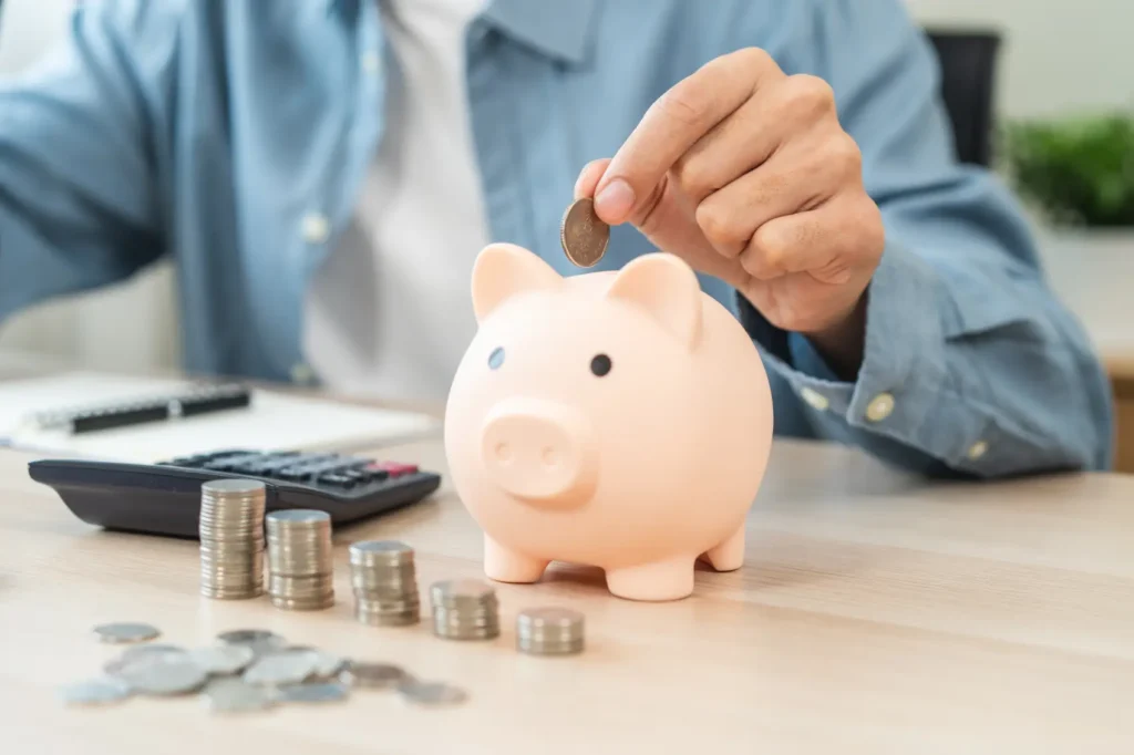 investimentos-seguros Jovem mulher economizando dinheiro com despesas mensais colocando moeda em um cofrinho na mesa.