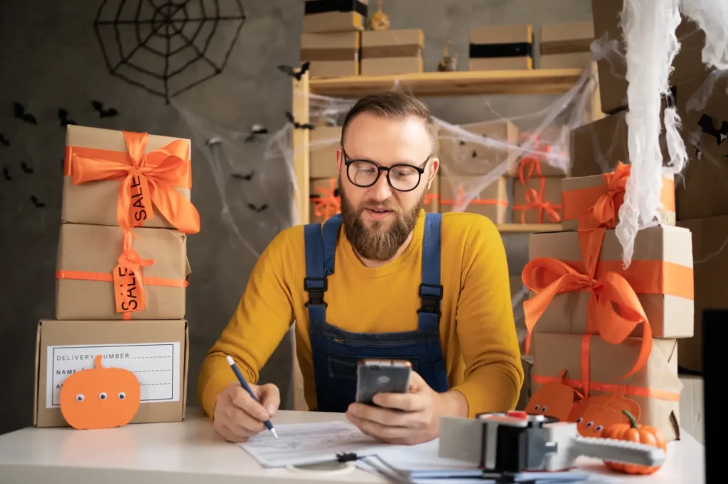 ideias de halloween - homem branco escrevendo em caderno, vestindo avental azul e caixas de estoque com decoração de hallowwen ao fundo