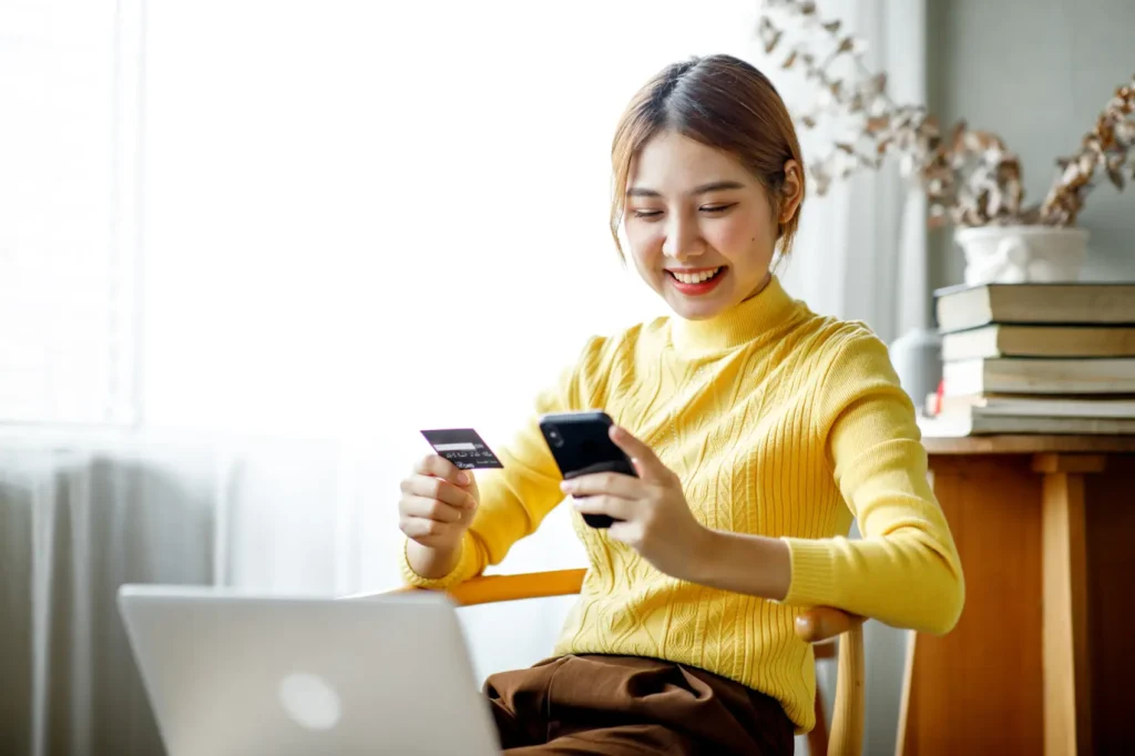 o que é conta corrente - Jovem asiática usando smartphone e notebook, segurando um cartão de crédito para fazer compras online