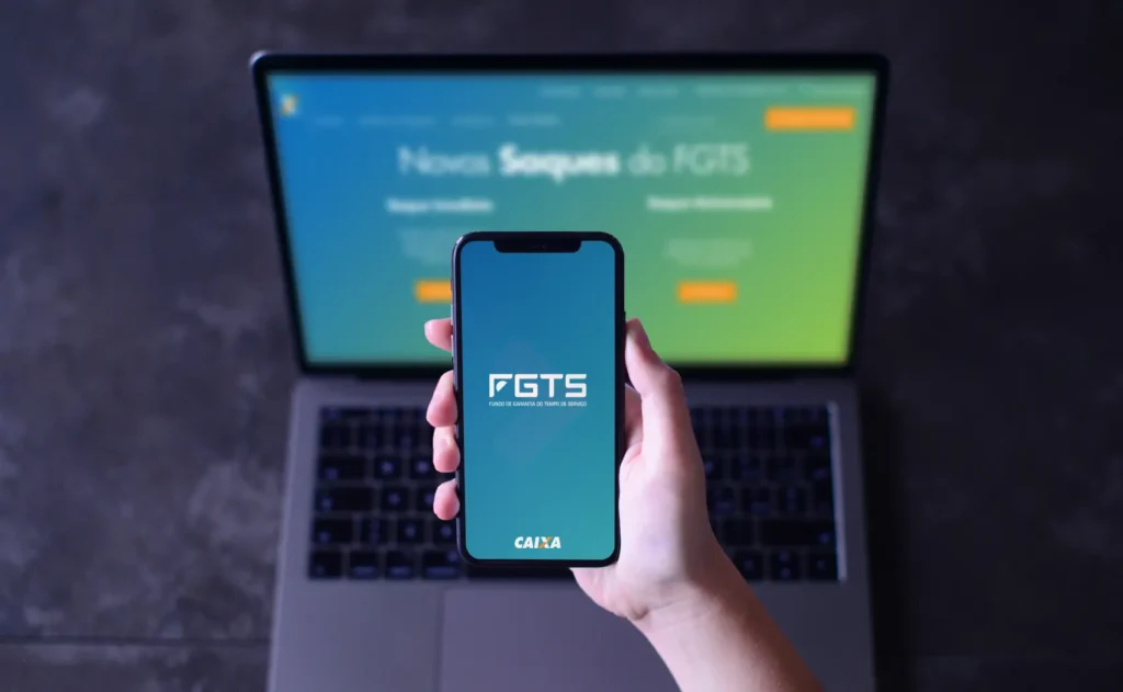 Saque-aniversário FGTS - Mão segurando um celular exibindo o aplicativo FGTS (Fundo de Garantia por Tempo de Serviço) da Caixa Econômica Federal (CEF) e um laptop mostrando a página de saque do FGTS