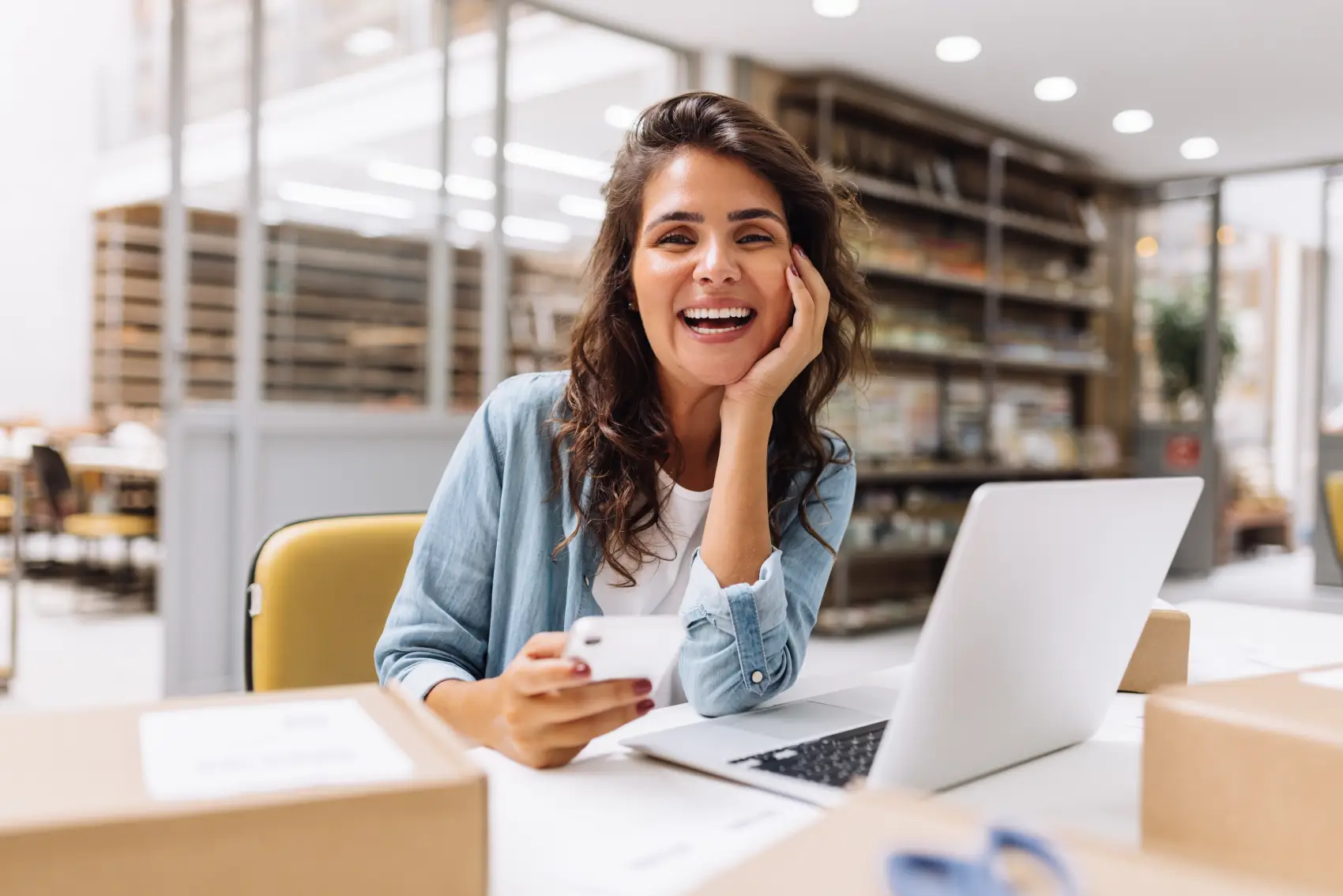 cdb com liquidez diária - mulher caucasiana sorrindo em frente ao notebook com celular na mão, olhando diretamente para a câmera