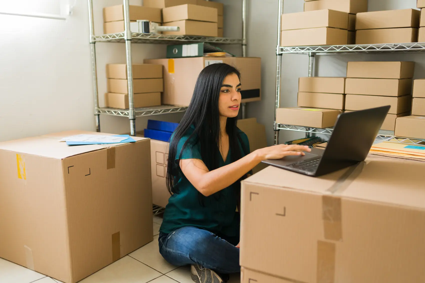 o que é controle de estoque - mulher sentada no chão, apoiando seu notebook em uma caixa, e com várias caixas ao seu redor