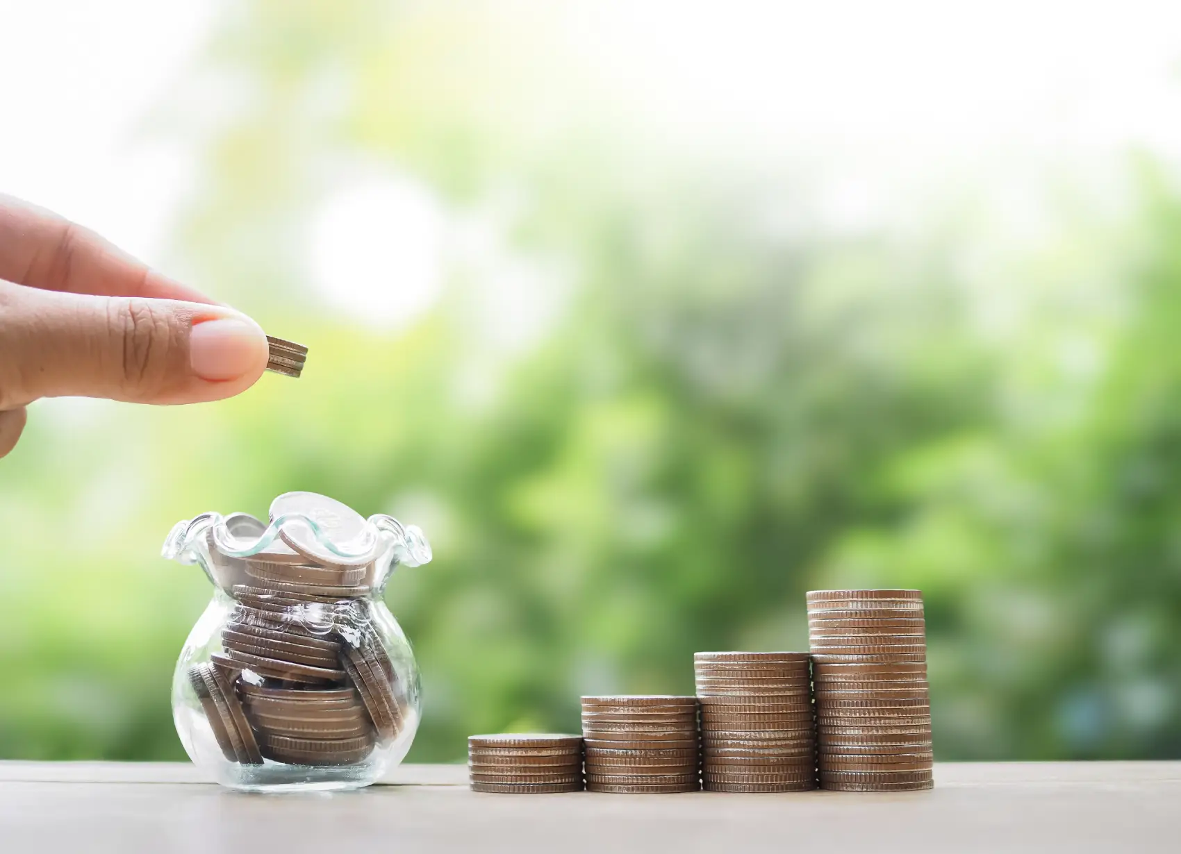 como investir com pouco dinheiro - Close de uma mão colocando uma moeda em uma garrafa de vidro e uma pilha de moedas