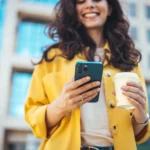 o que é conta digital - Jovem empresária de atitude positiva, com um copo de café para viagem na mão, usando o celular