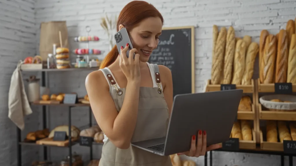 programa acredita​ - Jovem conversando ao telefone enquanto usa o laptop em uma padaria com pão e doces nas prateleiras ao fundo