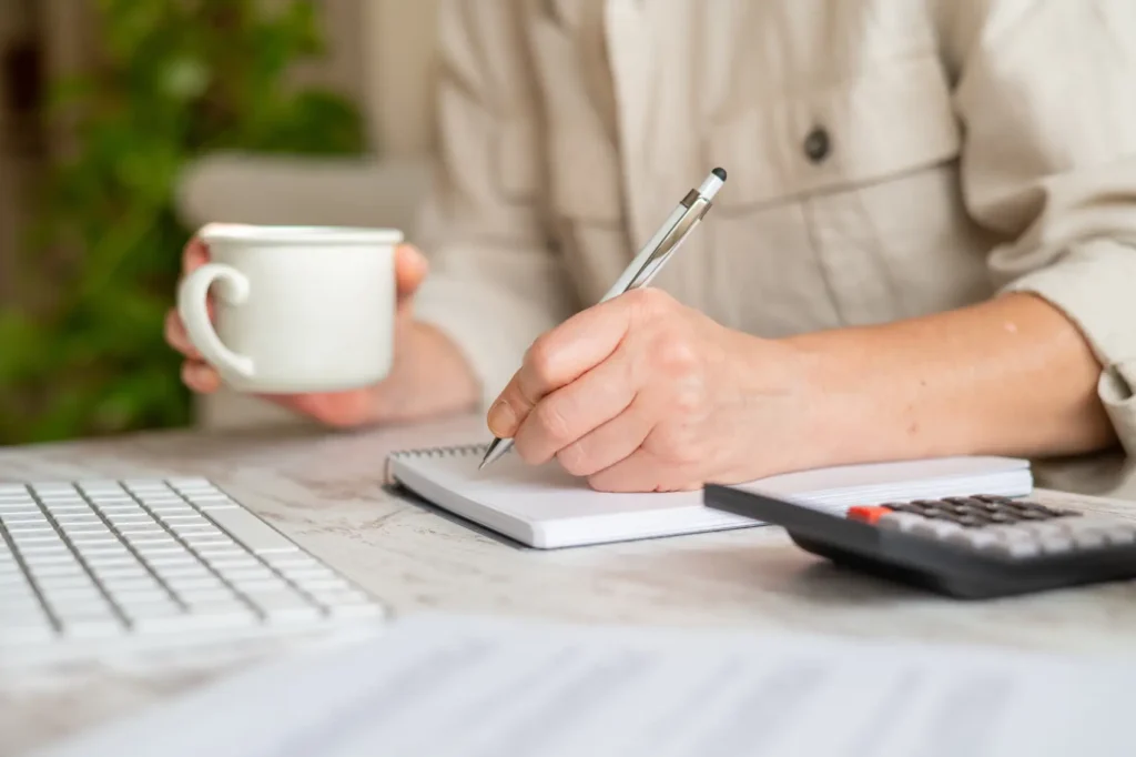 como economizar dinheiro - Mulher com foco em finanças pessoais, gerenciamento de despesas domésticas e avaliação de planilhas de orçamento enquanto escreve notas em um caderno espiral com uma calculadora e uma xícara de café
