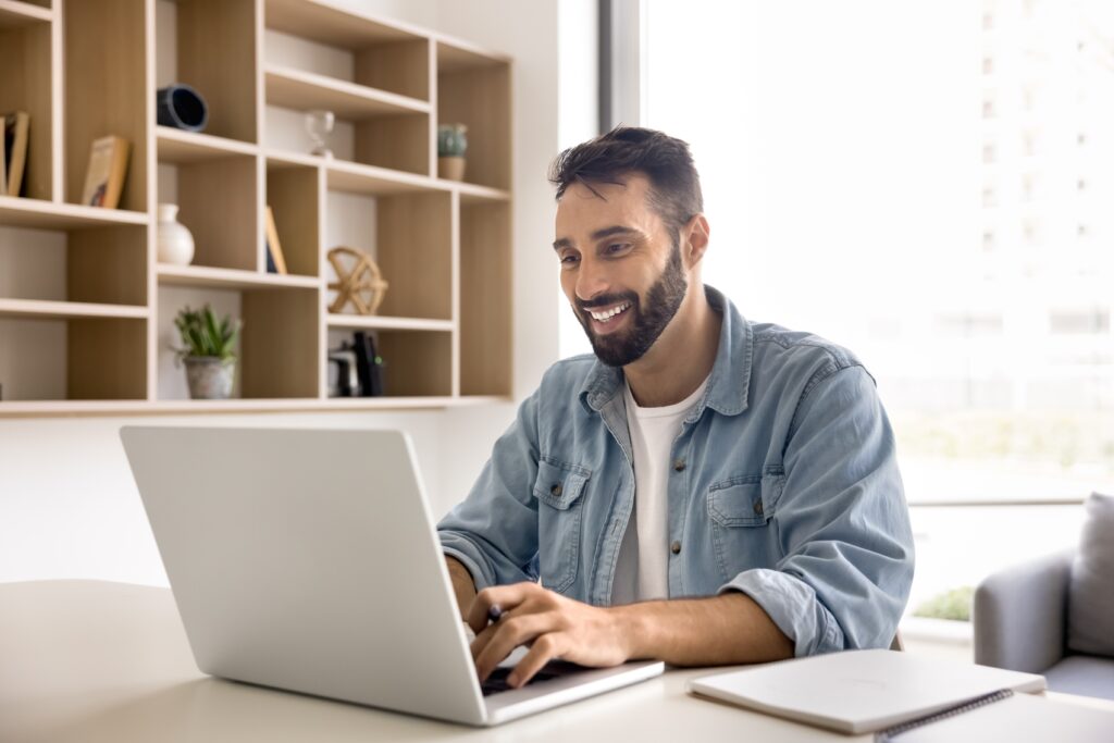 planilha de gastos​ - Jovem sorridente homem de negócios anotando informações úteis sentado na mesa com computador moderno
