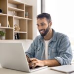 planilha de gastos​ - Jovem sorridente homem de negócios anotando informações úteis sentado na mesa com computador moderno