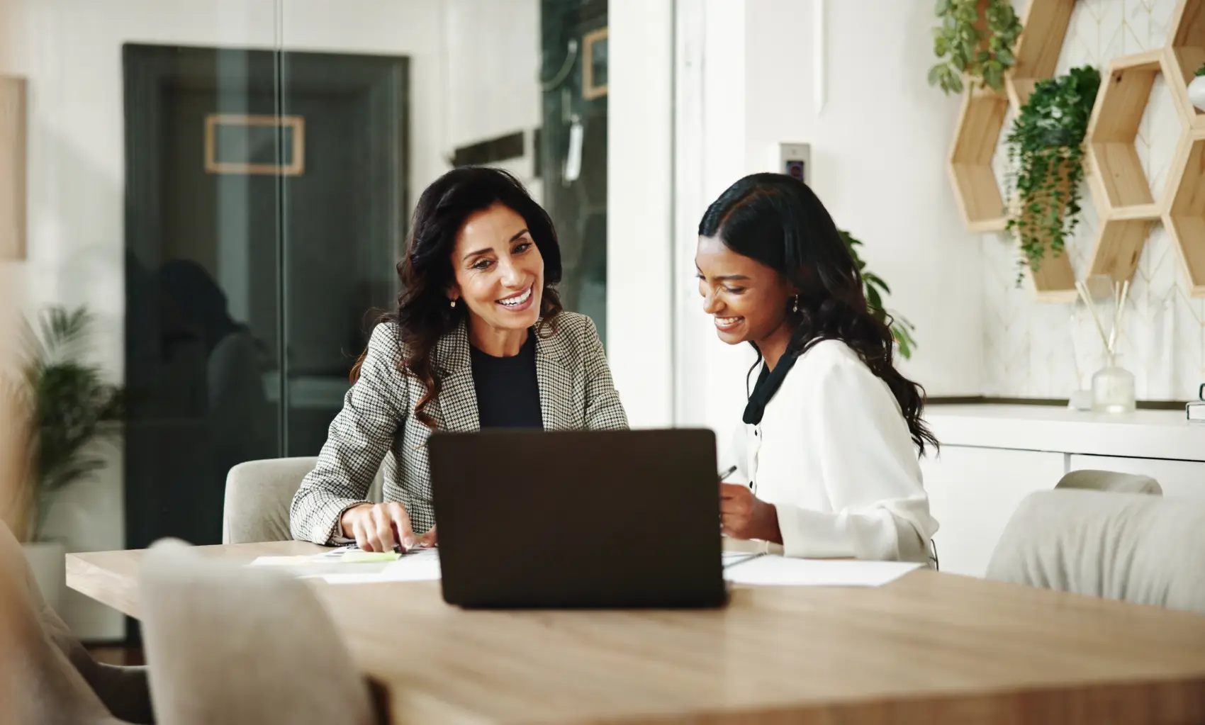 como investir em renda fixa - mulher branca e mulher preta sorrindo olhando para o notebook, sentadas em frente a mesa de madeira