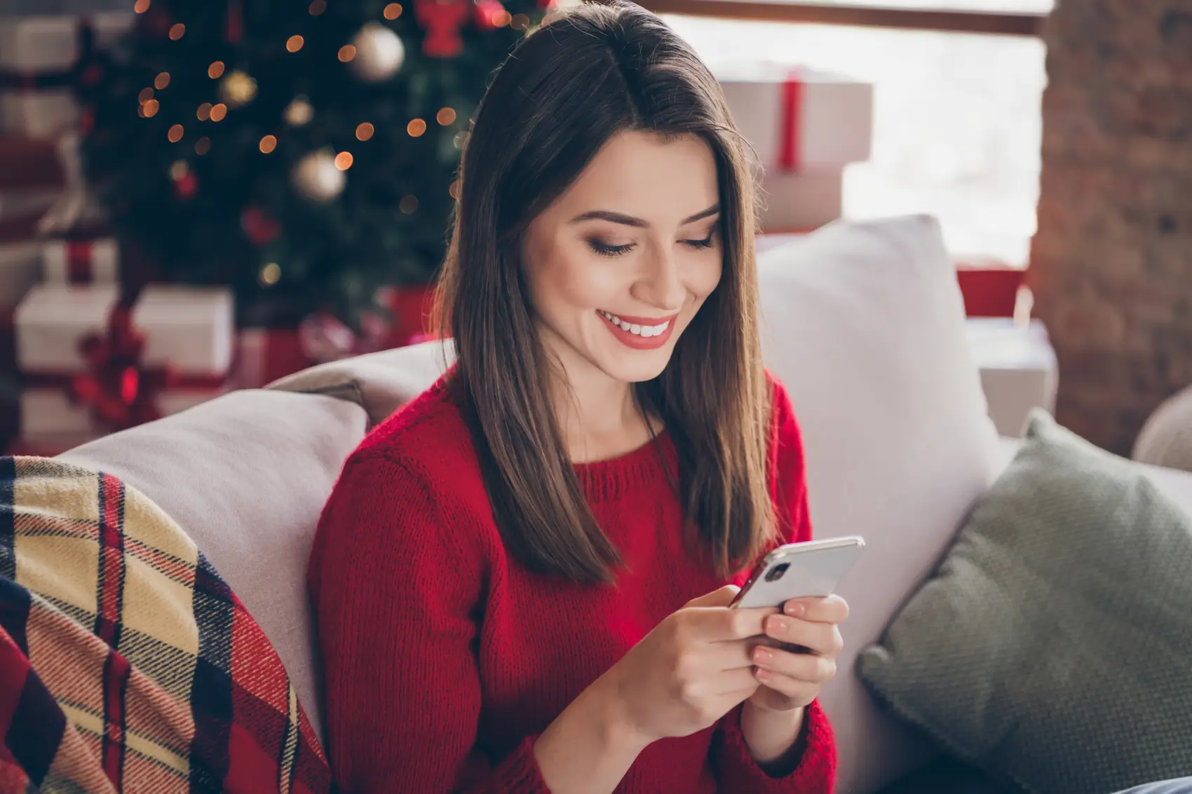 mensagem de natal para clientes - Foto de menina sorridente segurando o smartphone, sentada confortavelmente em um sofá dentro de casa, com uma árvore de Natal atrás dela