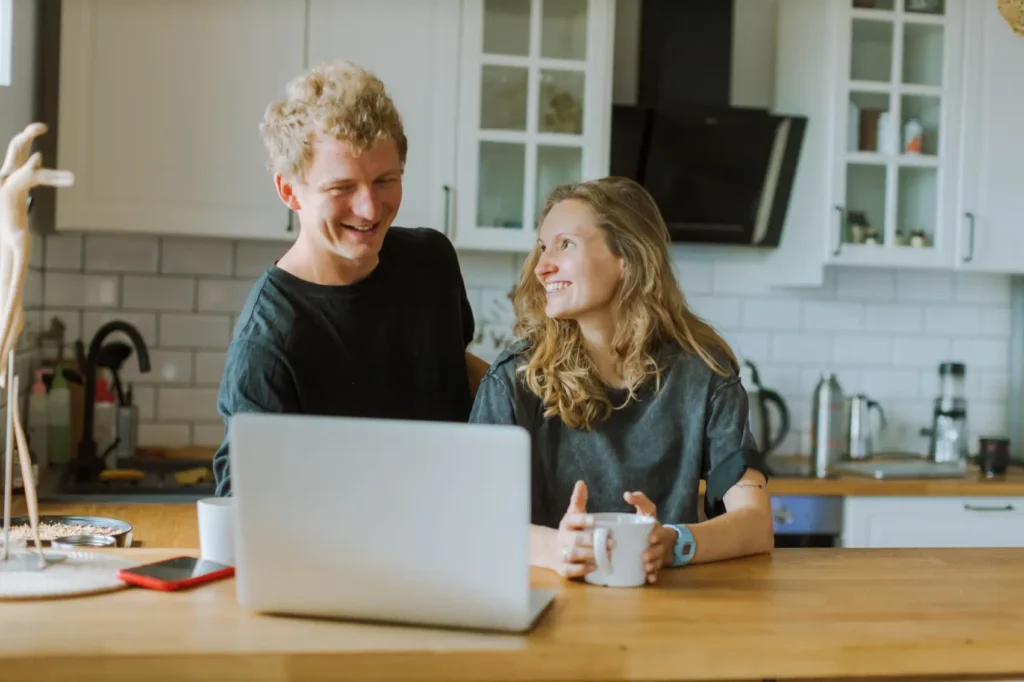 tudo-sobre-portabilidade-de-consignado Um casal alegre compartilha um café e risadas em sua cozinha com os olhares na tela de notebook.