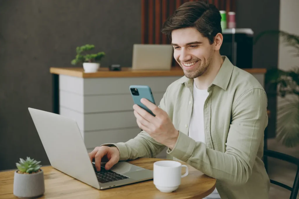 mudanças no fgts​ - homem sentado em frente ao notebook, olhando para o celular em sua mão e sorrindo