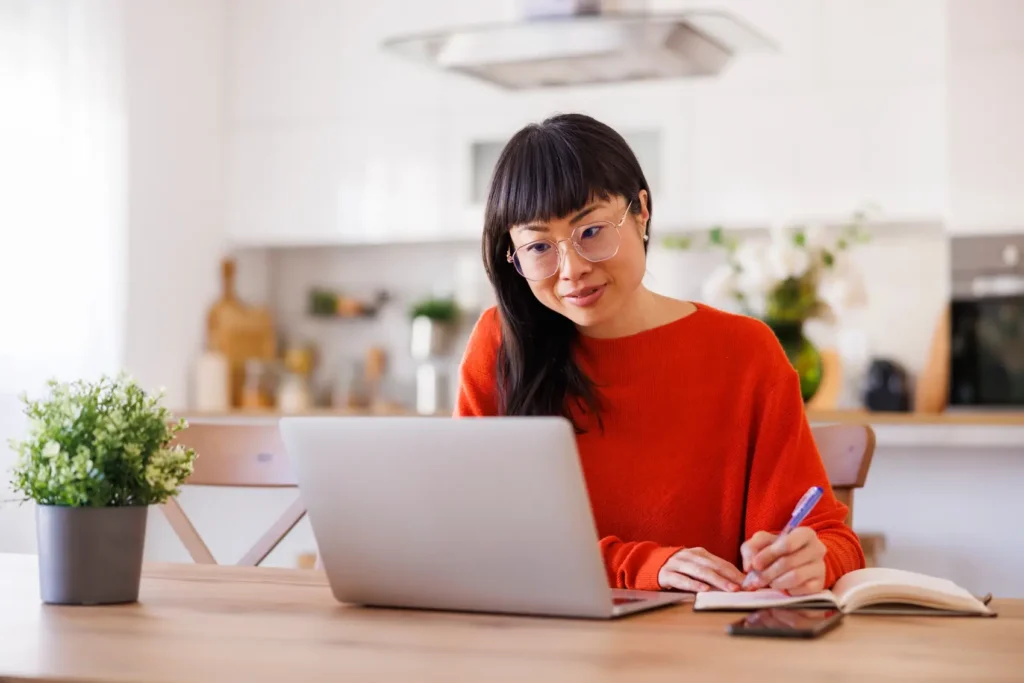 qual valor minimo para declarar imposto de renda - Mulher asiática madura anotando em sua agenda enquanto faz um curso online em casa usando um laptop
