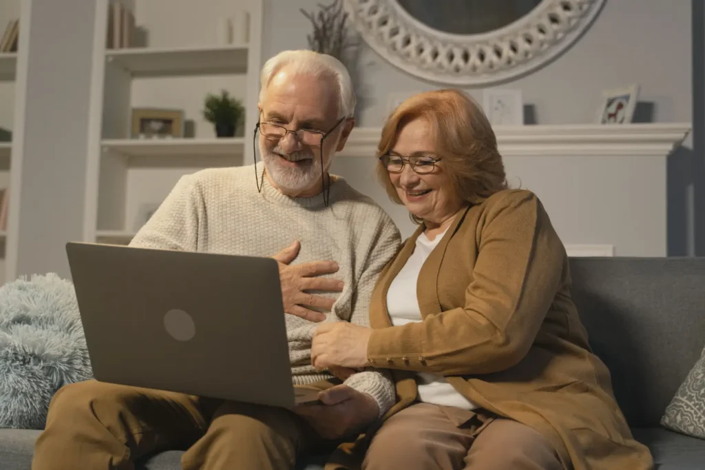 como-fazer-emprestimo-consignado Casal de idosos feliz sentado em um sofá fazendo uma chamada de vídeo alegre em um laptop em uma sala de estar aconchegante.