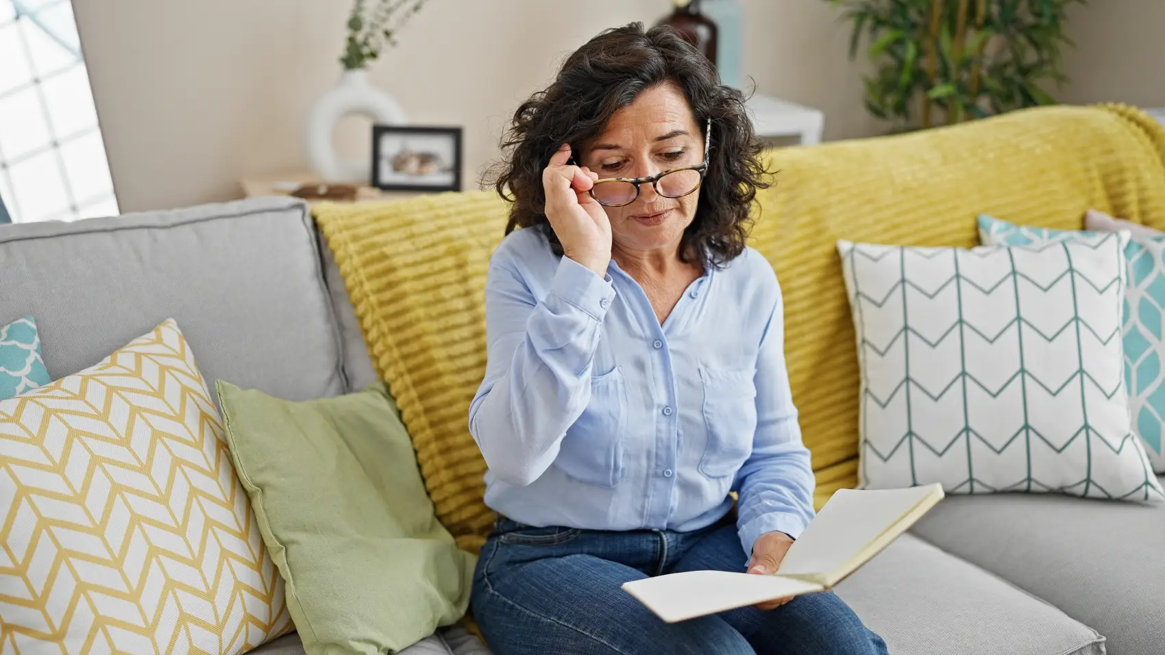 livros-sobre-investimentos Mulher hispânica de meia-idade lendo um livro sentada no sofá em casa