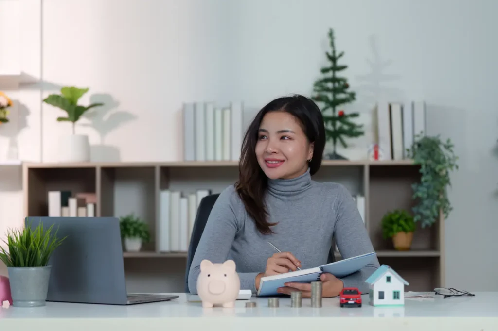 o-que-e-educacao-financeira mulher analisando finanças com cofre de porquinho, casa e carro em miniaturas