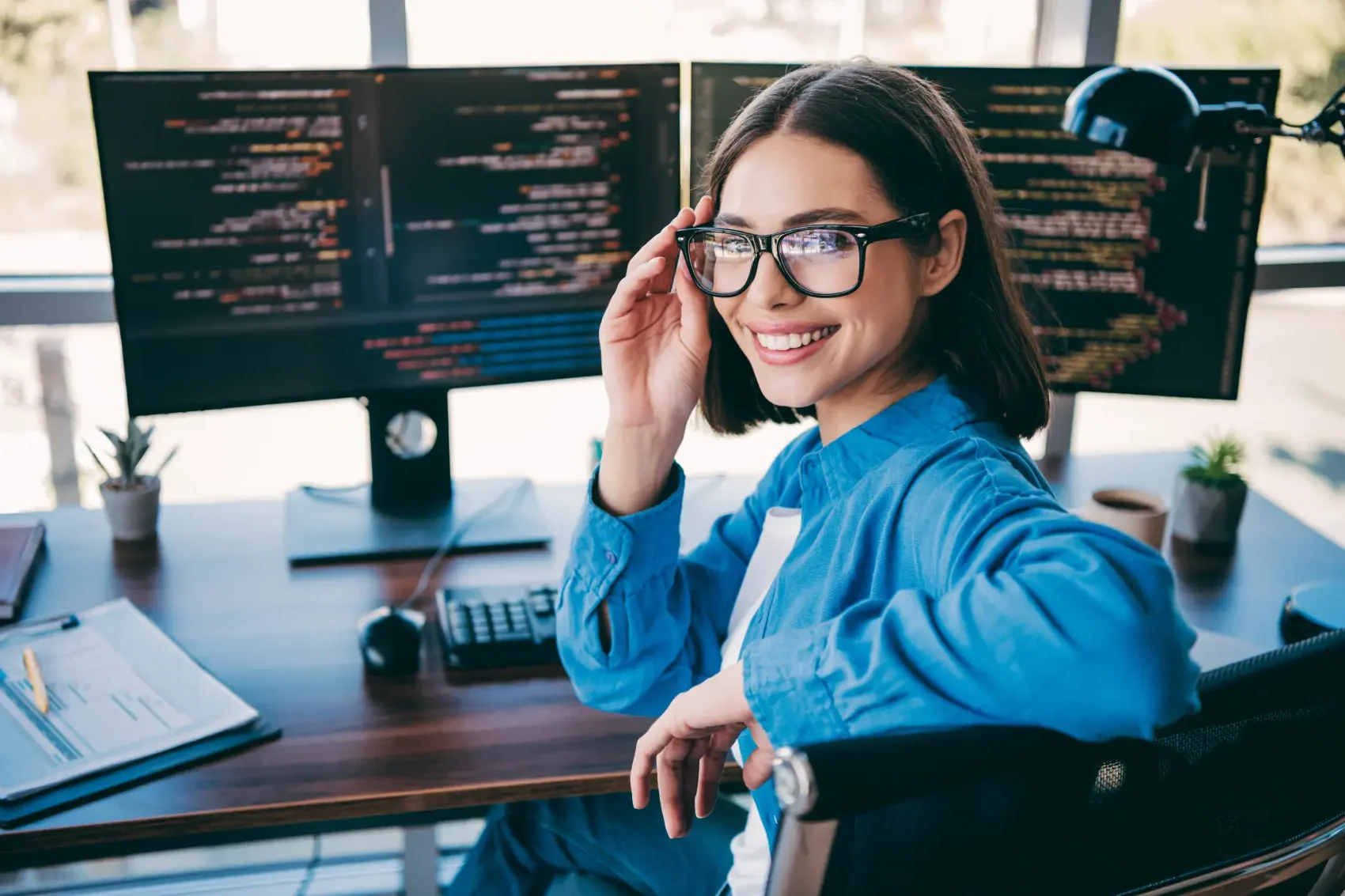 Como ganhar dinheiro na internet - Uma programadora alegre usando óculos senta-se em um espaço de trabalho limpo com monitores duplos mostrando código, um teclado e notebook nas proximidades, transmitindo foco, criatividade e trabalho técnico profissional em um escritório moderno