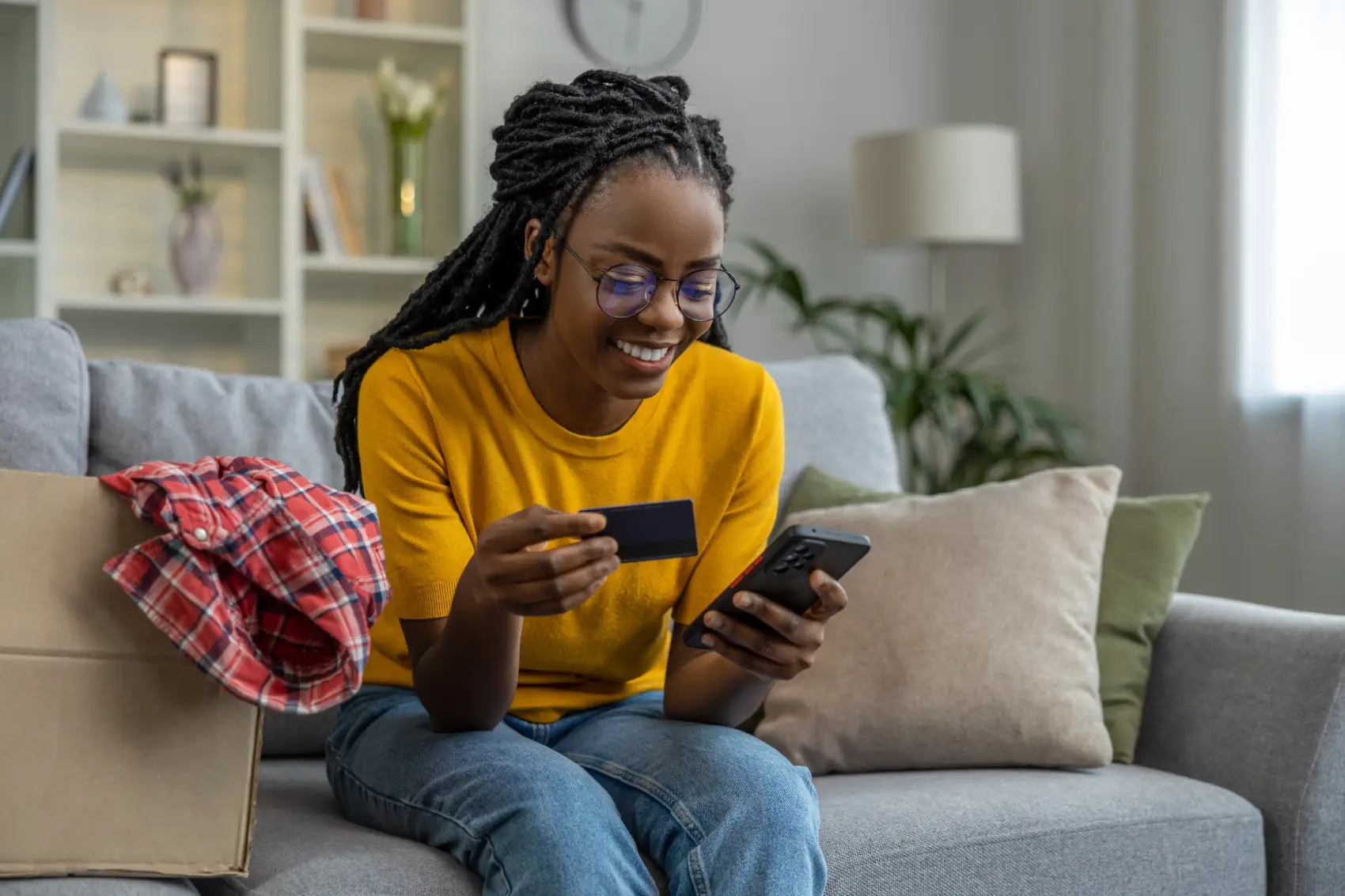 limite do cartão de crédito​ - mulher preta sentada em sofá bege sorri em frente ao celular, segurando um cartão de crédito preto