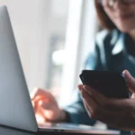 black fraude​ - pessoa sentada em mesa, em frente ao notebook e olhando o celular