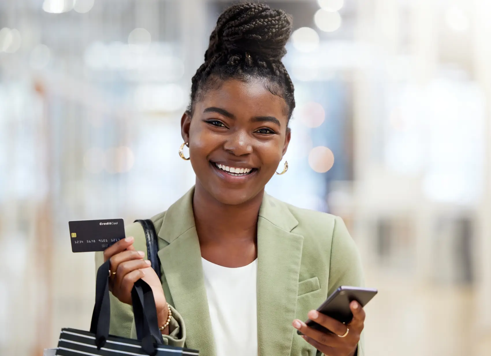 como funciona o parcelamento no cartão de crédito​ - mulher negra segurando cartão de crédito e sacola de compras em uma mão e celular na outra. ela sorri para a câmera com os cabelos presos em um coque alto