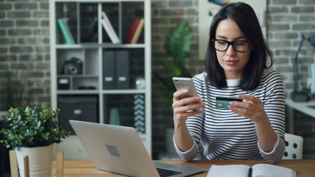 estorno-de-cartao-de-credito mulher com cartão de crédito e celular na mão em mesa de escritório com notebook