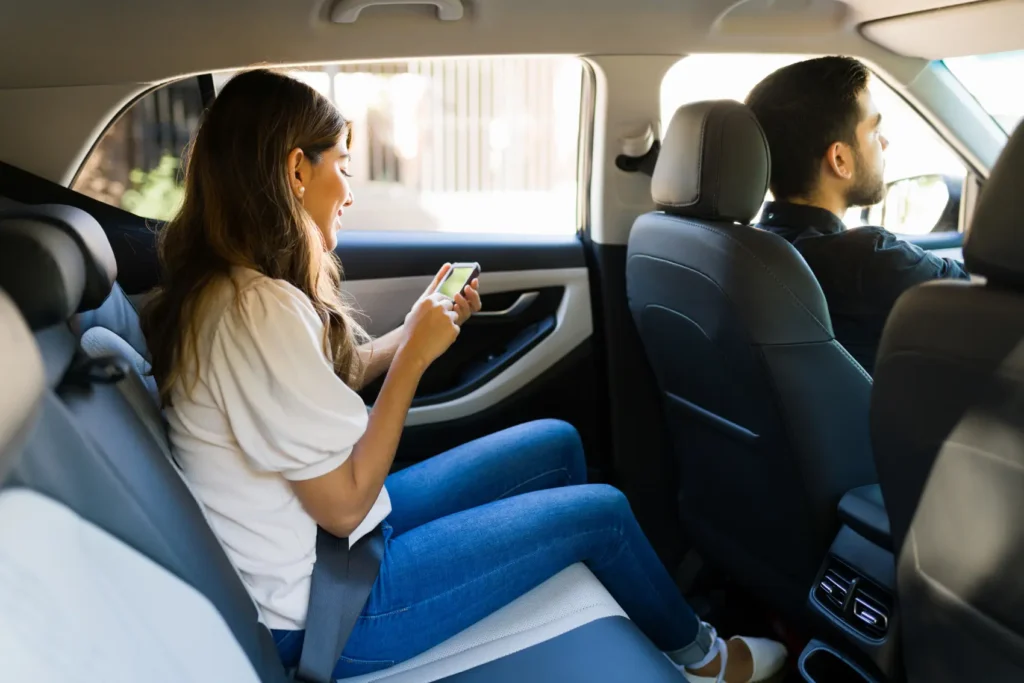 como ser motorista de aplicativo - Vista lateral de uma mulher latina atraente sentada no banco de trás de um carro, usando um aplicativo de transporte por aplicativo em seu smartphone