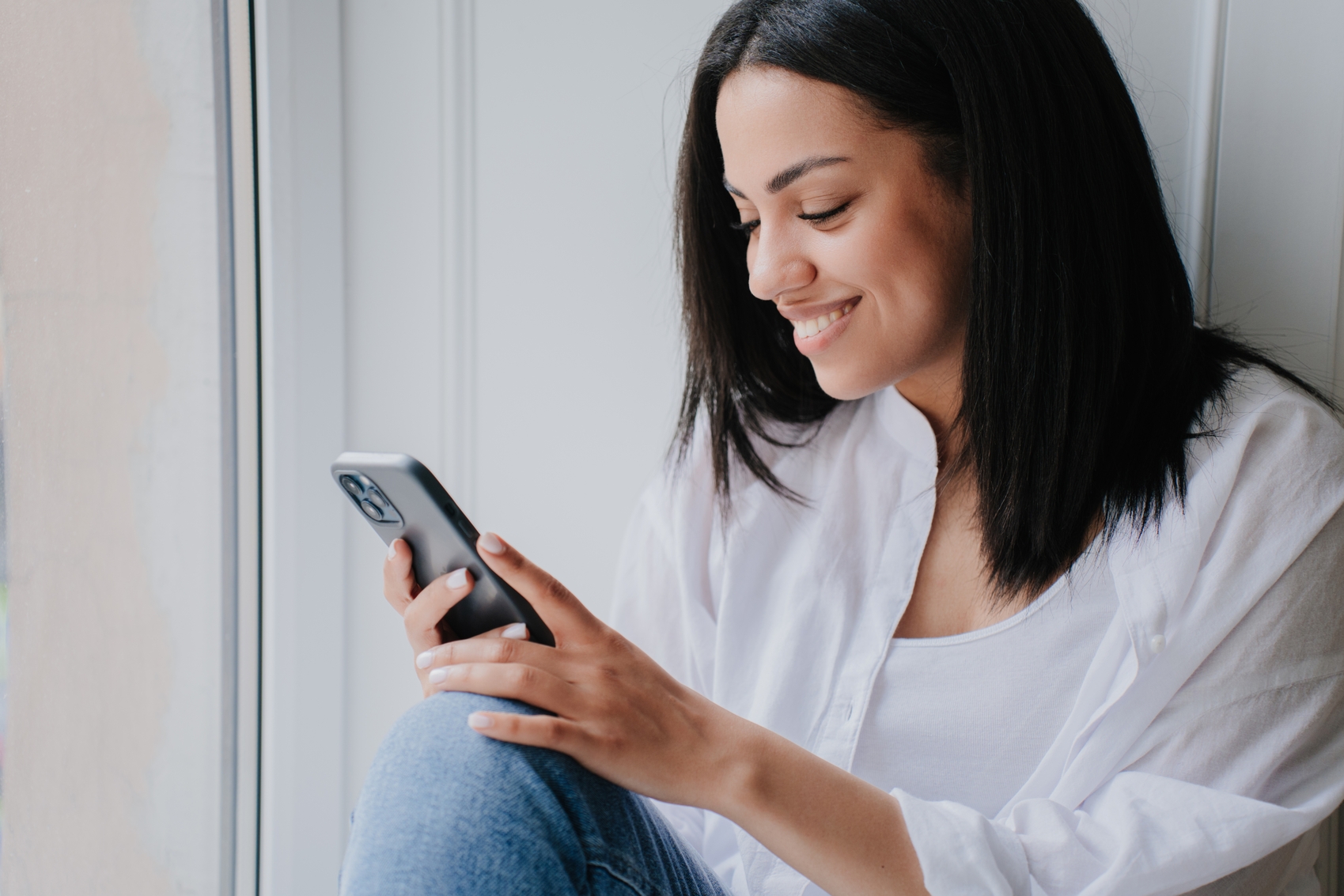 novas regras do imposto de renda - Alegre e jovem brasileira de camisa branca e calça jeans, segurando o celular com um sorriso radiante, olha para a tela após receber ótimas notícias