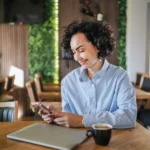 clt pode ser mei​ - Mulher com cabelo encaracolado sentada em uma mesa de madeira em um café moderno, verificando seu smartphone enquanto toma café expresso, laptop perto contra uma parede de planta verde,