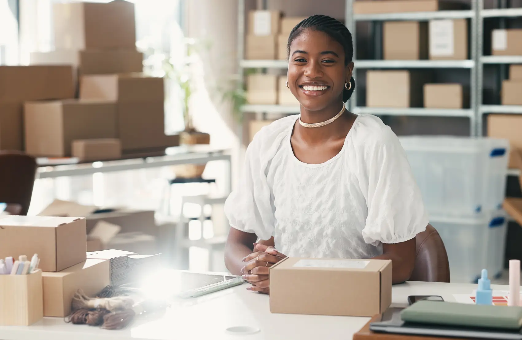 dia das micro pequenas e médias empresas - mulher nega sorrindo sentada em frente à mesa com caixas de estoque ao fundo