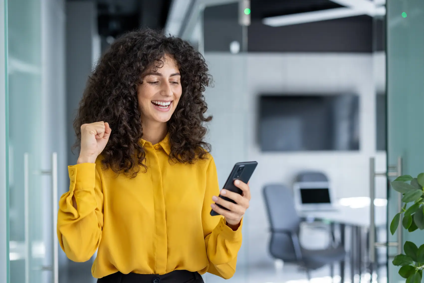crédito do trabalhador pagbank - Mulher alegre no escritório celebra o sucesso com o smartphone. De cabelos encaracolados em camisa amarela, ela sorri enquanto segura o telefone, expressando conquista