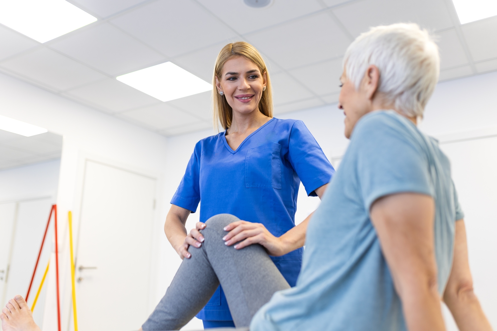 fisioterapeuta pode ser mei - Fisioterapeuta feminina sorridente apoiando uma mulher idosa durante exercícios de reabilitação do joelho em uma clínica moderna enquanto trabalham juntos para melhorar a mobilidade e a força após uma lesão